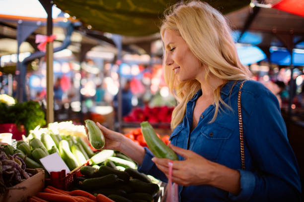 Mid adult woman buying vegetables on greenmarket. Choosing zucchini and placing in plastic bag.