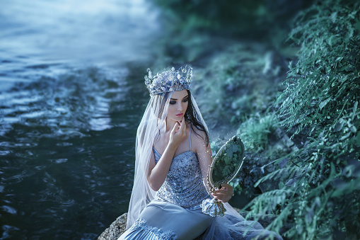 Snow White. The young lady looks in the mirror. The girl in a luxurious silver dress. Creative colors.