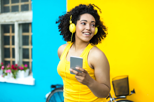 Smiling african woman with headphones and a bike using smart phone in front of blue and yellow wall backgound.