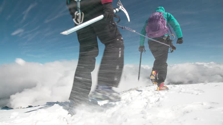 Two female mountaineers traverse a mountain ridge on a wind swept glacier at high altitude above the clouds.