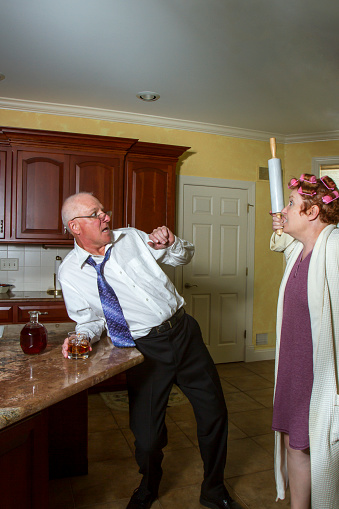 Drunk man leaning against counter in kitchen with angry wife in bathrobe and curlers threatening with rolling pin.