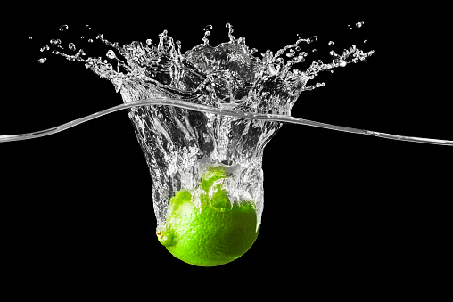 One fresh green juicy lime is falling into crystal clear water. There are bubbles surrounding the fruit and splashes of water rising high above the water surface from where the lime has broken the surface. The background is black.