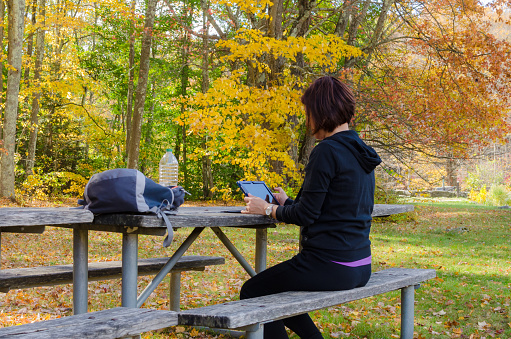 Woman in Tracksuit Surfing the net with her Tablet while Sitting on a Picnic Table with Colourful Autumnal Trees in Background