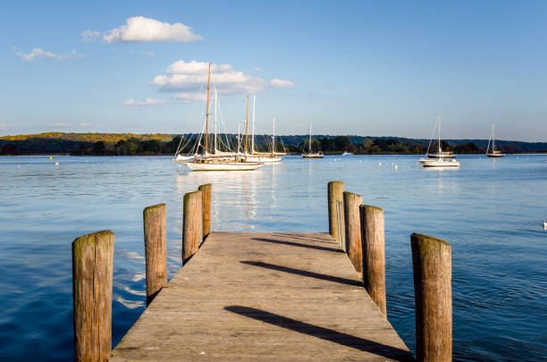 Empty Wooden Jetty on the Connecticut River at Sunset. Some Sailing Boats are anchored in fornt of the Jetty. The Forested Bank of the River is Visible in Background. Essex, CT.