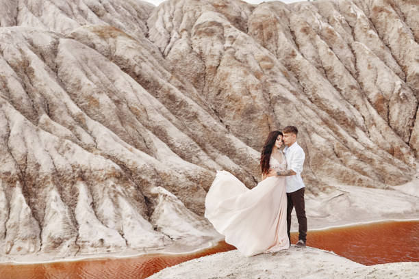 couple stands on the precipice of the mountain and the red lake and hugging. the bride and groom in love, the woman in long dress in the arms of men - long hair red hair women men imagens e fotografias de stock