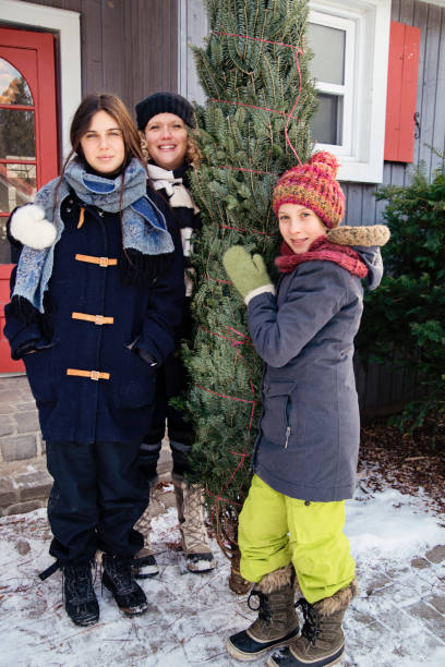 Family portrait with freshly cut Christmas tree in front of house outdoors. Family portrait with freshly cut Christmas tree in front of house outdoors. There is a bit of fresh snow on the ground. Family of mother, and two teenage girls. Vertical full length outdoors shot with copy space. This was taken in Quebec, Canada. 12 17 months stock pictures, royalty-free photos & images