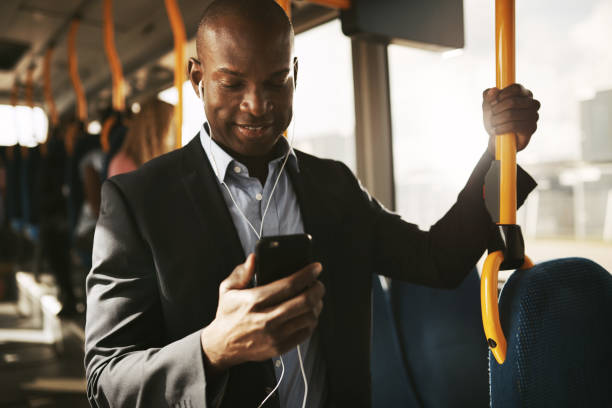 Young African businessman riding on a bus listening to music Smiling young African businessman wearing a suit standing on a bus during his morning commute listening to music on a smartphone and earphones commuter stock pictures, royalty-free photos & images