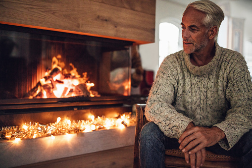 Mature man sitting by fire place in living room at home. Senior male relaxing on chair.
