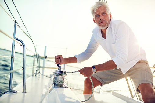 Mature man standing on the deck of a boat winding a winch while out for a sail on a sunny afternoon