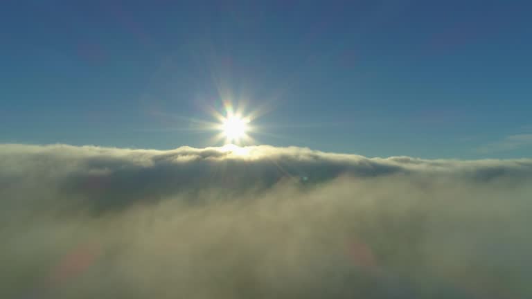 AERIAL: Flying above thick fog and clouds towards the golden sun on blue sky