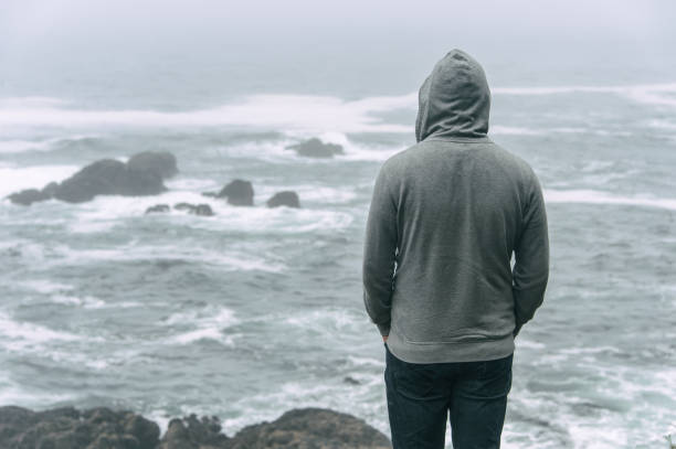 uomo triste e solitario in piedi di fronte all'oceano pacifico. - uomo incappucciato foto e immagini stock