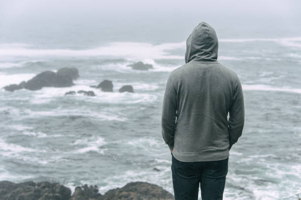 Sad and lonely man standing in front of the pacific ocean and looking at the stormy water on Vancouver Island.
