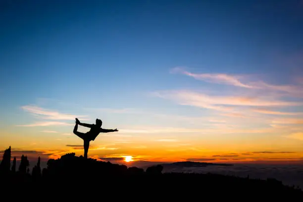 Woman meditating in yoga dancer(Natarajasana) pose silhouette over ocean and islands, coastline and rocky mountains. Inspiration and  motivation with healthy lifestyle in beautiful sunset landscape, fitness concept.