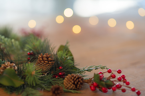 Close up shot of festive winter decorations. A Christmas wreath with red berries and pine cones lies on a rustic wood table. White Christmas lights twinkle in the background. Copy space is available in the upper right of the frame.