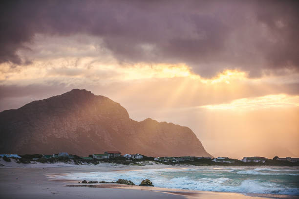 A dramatic cloudy sunrise taken from the beach over Bettys Bay Kleinmond Cape Town South Africa with the town in the foreground and warm sunbeams and clouds in the sky and a Kogelberg mountain background towards the nature reserve