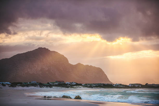 Sunrise over Betty's Bay A dramatic cloudy sunrise taken from the beach over Bettys Bay Kleinmond Cape Town South Africa with the town in the foreground and warm sunbeams and clouds in the sky and a Kogelberg mountain background towards the nature reserve hermanus stock pictures, royalty-free photos & images