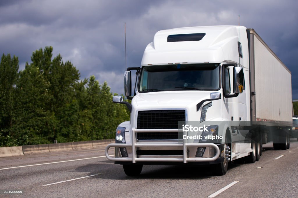 Big rig white semi truck with grille guard and trailer on the road A modern large white big rig semi truck tractor with a powerful grille protection and a spacious cabin and dry van semi trailer delivers the industrial cargo for long distances traveling on America's highways Semi-Truck Stock Photo