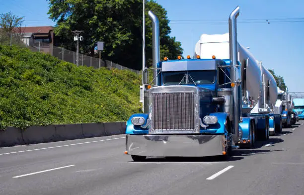Icon of style. A classic American bonneted big rig semi truck with high tailpipes and chrome parts majestically pulls the trailer for concentrated loads and rides ahead of the convoy of other trucks with various types of semi trucks and trailers and goods carried along the highway