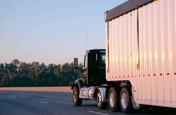 A modern, dark, compact, powerful big rig semi truck with a day cab for shorter journeys within a day's journey with a long semi trailer for the carriage of bulk cargo is moving along a wide highway illuminated by the warm rays of the evening sunset