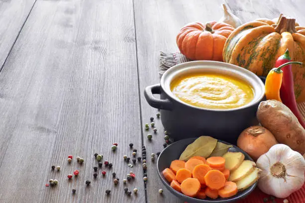 Pumpkin, carrot and ginger creme soup in a dark ceramic pan on the wooden table. Ingredients: pumpkins, carrots, ginger, onions, garlic, chili, salt and pepper. Text space.