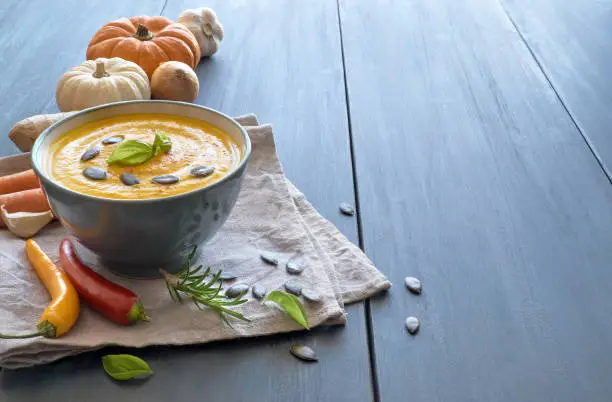 Spicy pumpkin creme soup with carrot and ginger seasoned with pumpkin seeds and basilicum. Ingredients: pumpkins, carrots, ginger, onions, garlic and chili. Soup is in ceramic bowl on blue wood, text space.