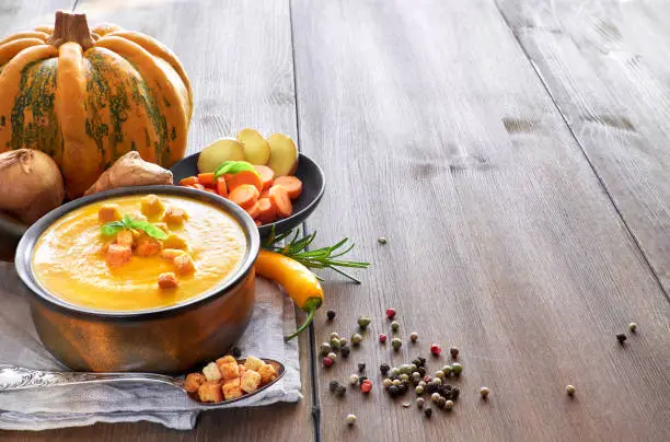 Pumpkin, carrot and ginger creme soup in ceramic pan on the wooden table, seasoned with croutons and basilicum. Ingredients: pumpkin, carrots, ginger, onions, cjili and colored pepper. Text space.