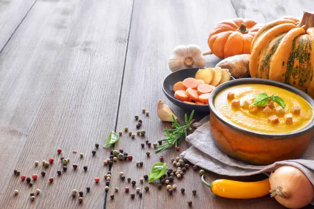 Pumpkin creme soup with carrot and ginger in ceramic bowl on wood, seasoned with croutons and basilicum. Ingredients: pumpkins, carrots, ginger, onions, garlic, chili and pepper. Text space.