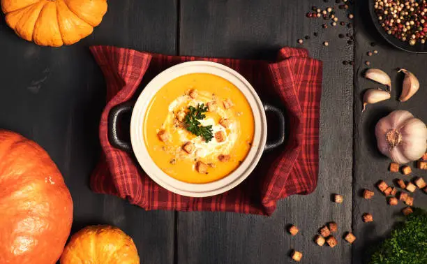 Pumpkin creme soup in a dark ceramic pan served with croutons, crushed nuts, parsley and cream, top view. Around are ingredients for the soup: pumpkins, garlic, parsley, croutons and colored pepper.