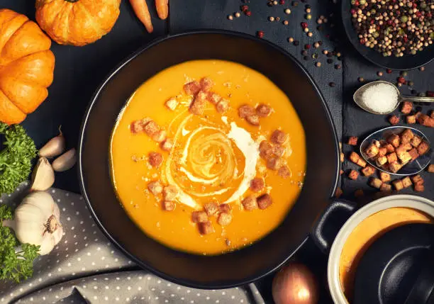 Pumpkin creme soup in a dark crockery served with croutons, crushed nuts and cream, top view. Around are ingredients: pumpkins, carrots, onions, garlic, parsley, colored pepper and salt.