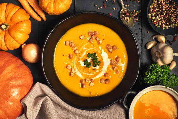 Pumpkin creme soup in a dark crockery served with croutons, crushed nuts and cream, top view. Around are ingredients: pumpkins, carrots, onions, garlic, parsley and  colored pepper.