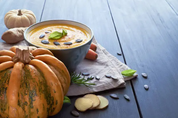 Spicy pumpkin creme soup with carrot and ginger seasoned with pumpkin seeds and basilicum. Ingredients: pumpkins, carrots, ginger, onions, garlic and chili. Soup is in ceramic bowl on blue wood, space