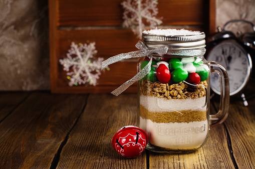 Cookies mix with color candies in a jar with paper tags. Handmade Christmas gift over dark stone background. Selective focus