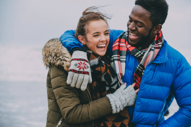 Young Couple Laughing On A Winter Beach A young couple with arms around each other laughing and having fun. teen romance stock pictures, royalty-free photos & images