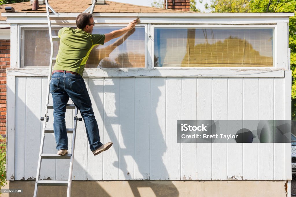 Man reaching to paint white trim on ladder White, middle aged male paints white trim of old urban home, and almost falls from ladder Ladder Stock Photo