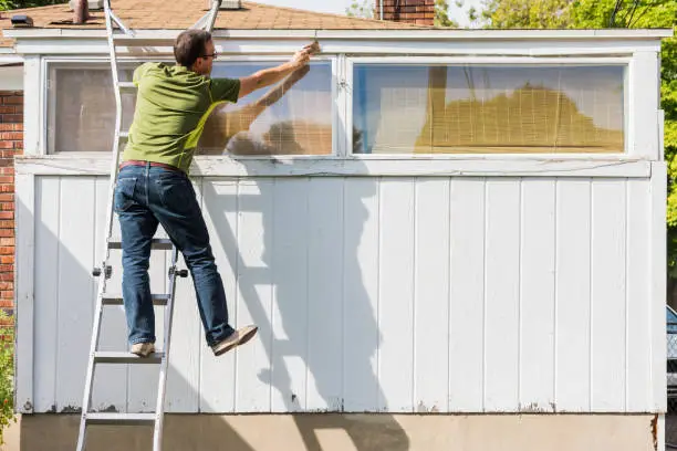 Photo of Man reaching to paint white trim on ladder