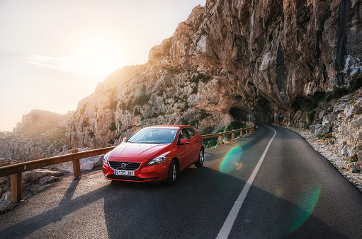Alcudia, Mallorca, Spain - May 24, 2016: Red car Volvo V40 traveling on the mountain serpentine through a tunnel of a rock along the coast of Majorca against the sunshine. Roadtrip around Spain