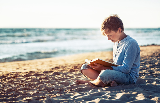 Cute boy reading a book on beach near sea at sunset