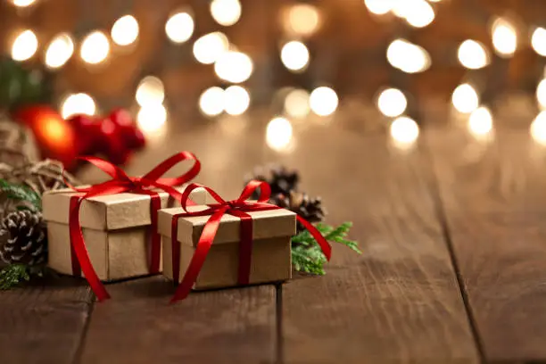 Horizontal view of two brown Christmas gift boxes shot on rustic wood table. The boxes are tied up with a red ribbon and Christmas lights are out of focus behind the group of boxes. The two boxes are grouped at the left side of the frame leaving a useful copy space ready for text and/or logo at the right. Predominant colors are brown and red. DSRL studio photo taken with Canon EOS 5D Mk II and Canon EF 70-200mm f/2.8L IS II USM Telephoto Zoom Lens