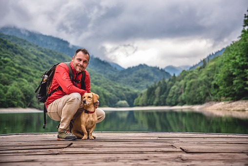 Hiker wearing red jacket and backpack with a small yellow dog on a pier posing by the mountain lake and foggy forest