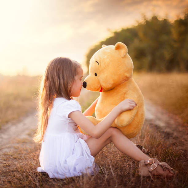 Little girl in white dress sitting on the ground in the meadow and playing with Teddy Bear at golden sunset.