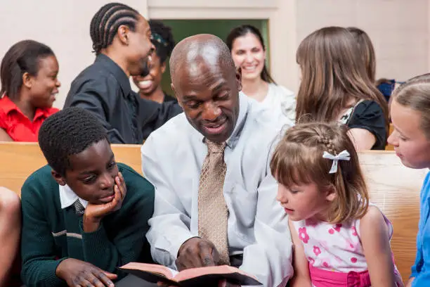 A multi-ethnic group of people are indoors in a church. They are wearing clothes suitable for church. A man of African descent is reading a bible to some children.