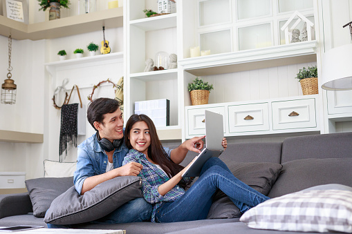 Young Asian couple relaxing together on sofa and using laptop computer at home in the living room