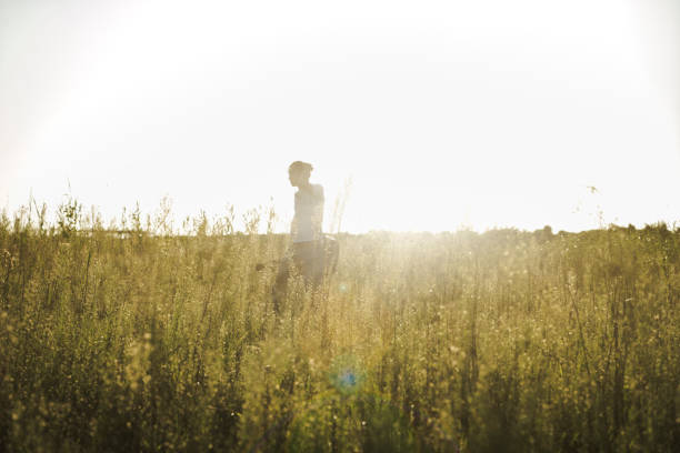 young asian man walking in field with guitar against sunbeam.