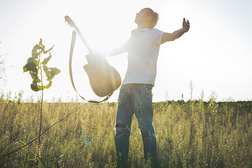 young asian man standing with arms raised in field holding guitar against sky.