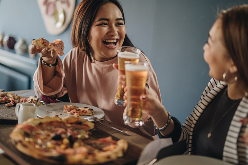 Happy Korean woman toasting with her friend while eating pizza in a restaurant.