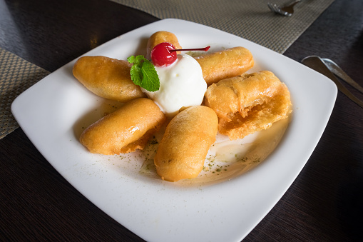 Fried banana fritters with vanilla ice cream, whip cream and cherry fruit on white plate on wooden table background