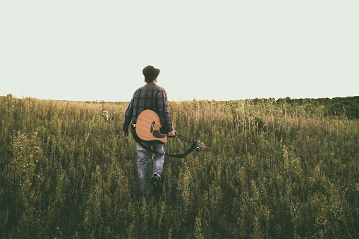 young asian man walking in field with guitar against sunbeam.