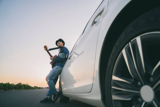 молодой мужчина, играющий на гитаре, опираясь на капот автомобиля - 20s acoustic guitar adult art стоковые фото и изображения