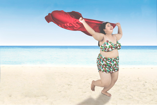 Joyful obese woman wearing bikini and running on the beach while holding a red scarf