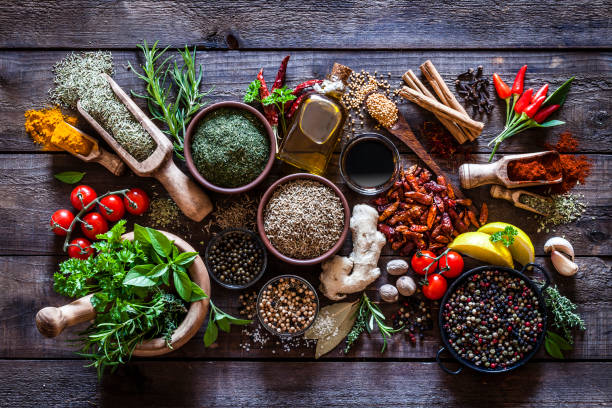 Spices and herbs on rustic wood kitchen table Top view of a rustic wood kitchen table filled with a large group of multi colored spices and herbs in bowls, wooden serving scoops or placed directly on the table. Spices and herb included are clove, turmeric, bay leaf, cinnamon, olive oil, curry powder, ginger, nutmeg, peppercorns, cinnamon, salt, chili pepper, basil, parsley, lemon, rosemary, garlic, onion and saffron. Low key DSRL studio photo taken with Canon EOS 5D Mk II and Canon EF 100mm f/2.8L Macro IS USM condiment stock pictures, royalty-free photos & images